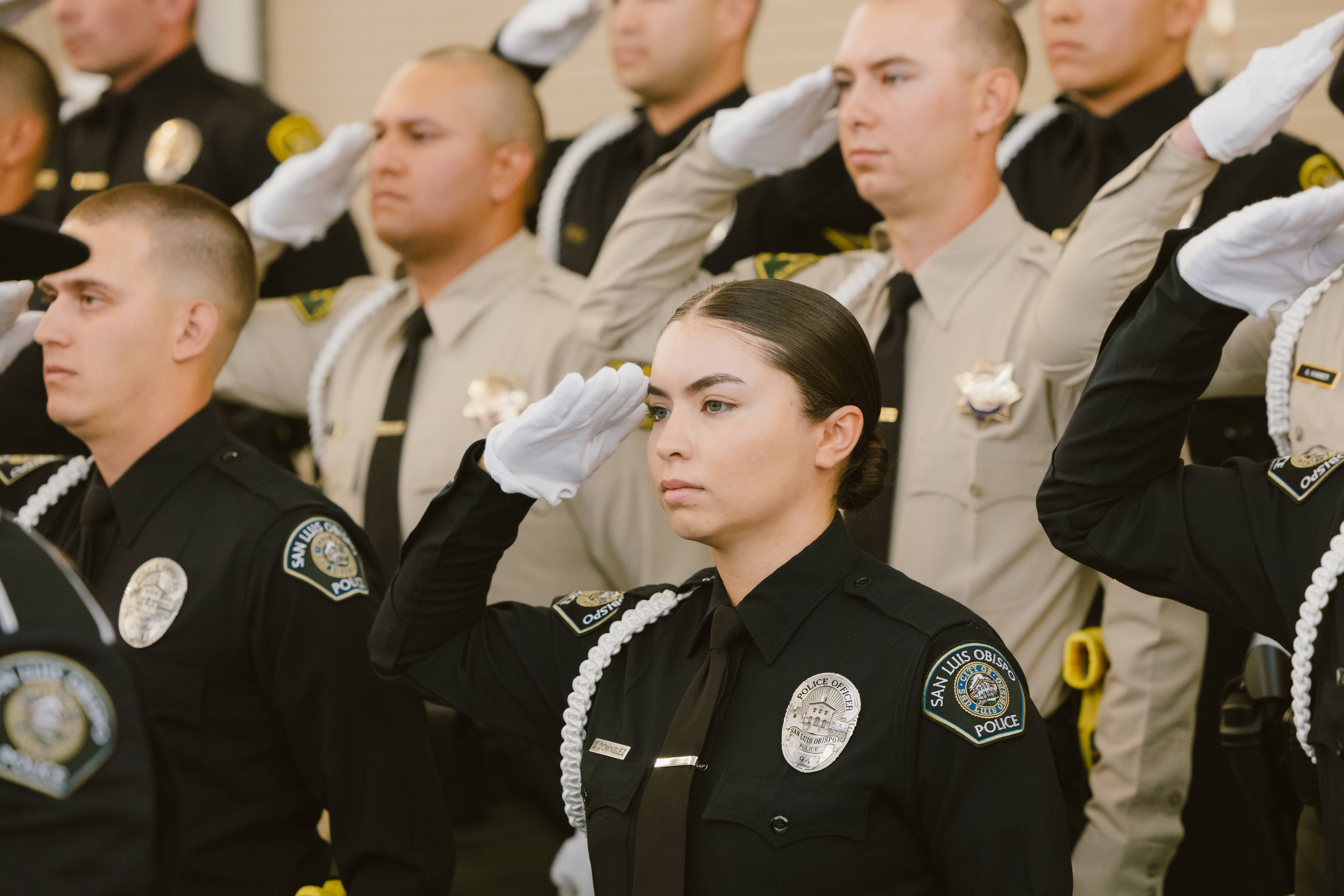 Law enforcement academy graduates stand in formation and salute during Allan Hancock College’s Law Enforcement Academy graduation ceremony. Recruits in police and sheriff uniforms wear white gloves and ceremonial cords as they participate in the formal oath and recognition proceedings.